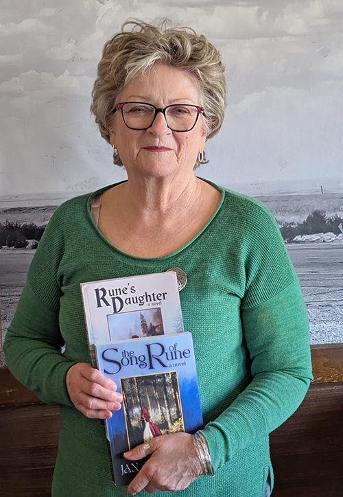 Photo of Jan Meadows standing indoors in front of an old photo mural, holding two of her fantasy series books.